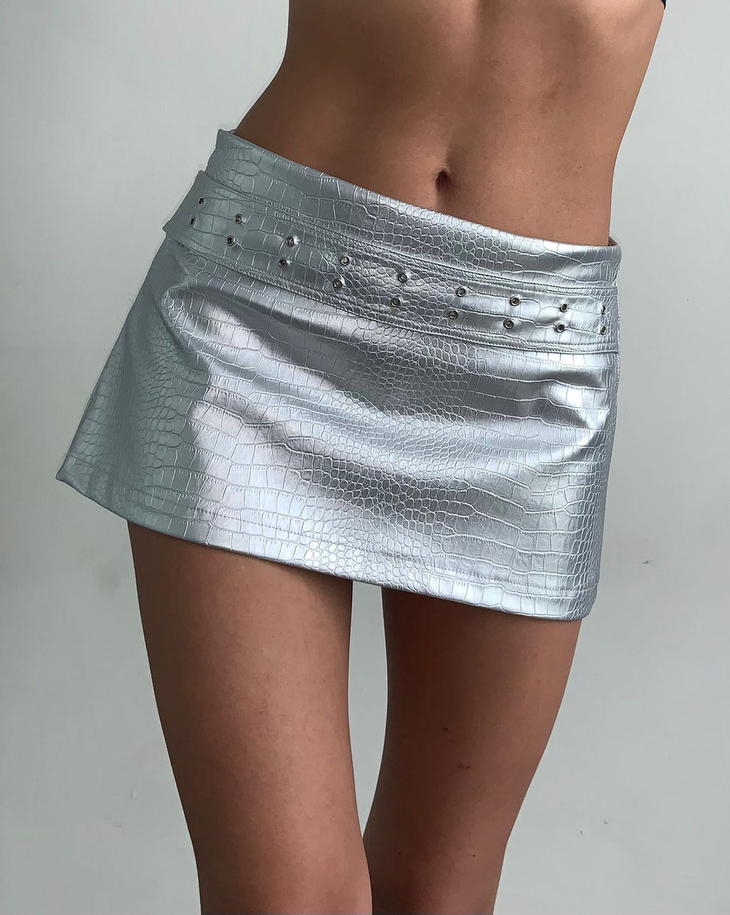 Mini falda confeccionada en cuerina con acabado metalizado, una tela que cede y se adapta al cuerpo, realzando la silueta sin perder estructura.

Cuenta con una correa de la misma cuerina, cosida directamente a la falda, que incorpora ojales metálicos, aportando un detalle moderno y funcional. Su silueta recta y corta le da un aire contemporáneo con una estética limpia y editorial.